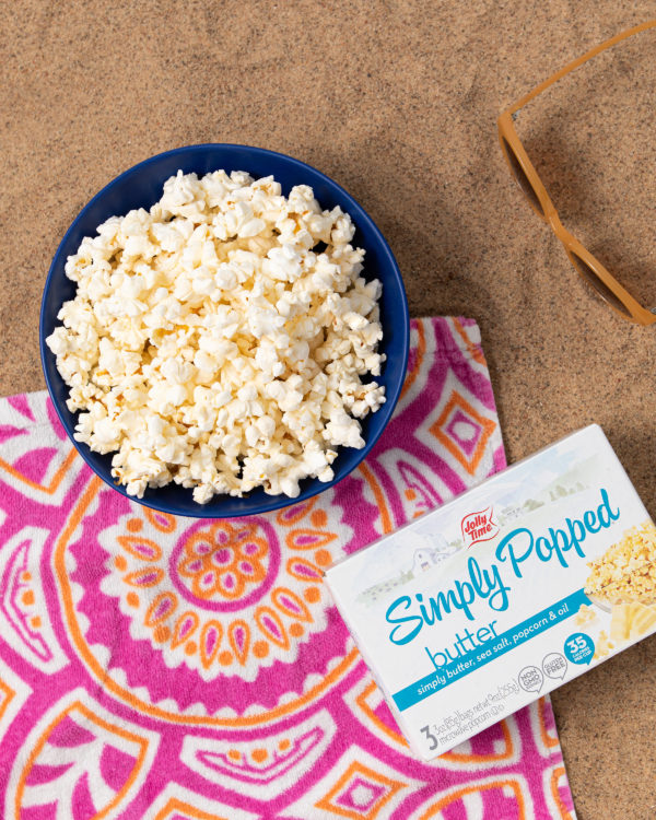 A bowl of popcorn laying on a sandy beach with a beach towel, sunglasses and a box of Simply Popped Butter microwave popcorn. 