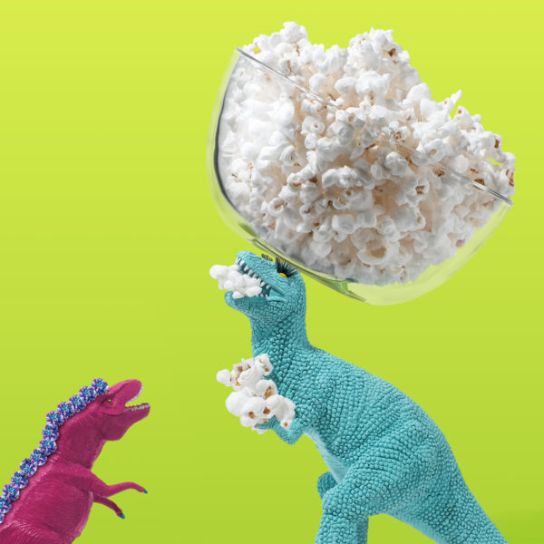 A photo of two toy dinosaurs, the on the right is holding popcorn kernels while balancing a bowl of popcorn on his head. 